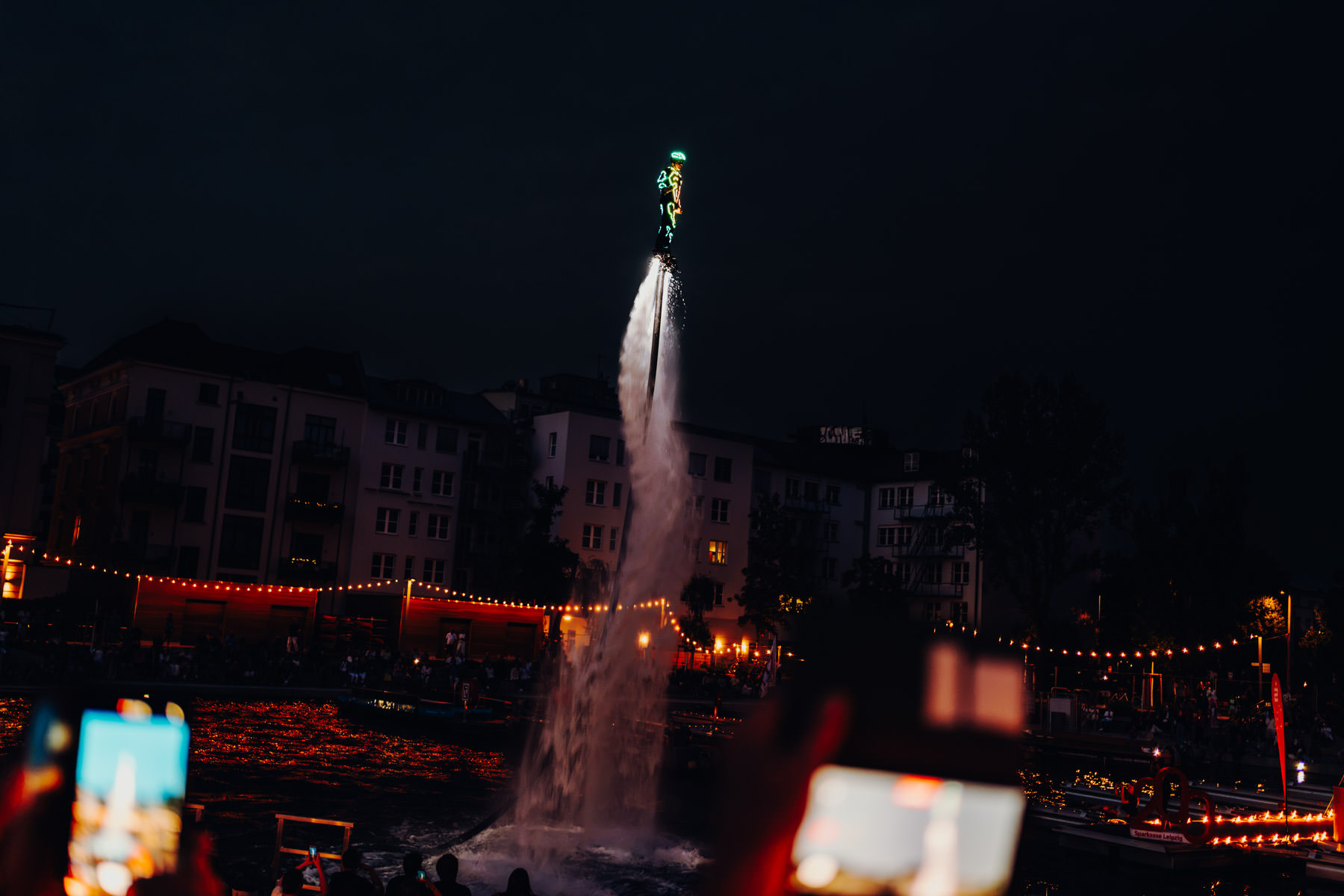 Eventfotograf Leipzig Flying Gordon Person auf einem Flyboard schwebt nachts auf einem hohen Wasserstrahl vor einer beleuchteten Stadtkulisse im Stadthafen Leipzig