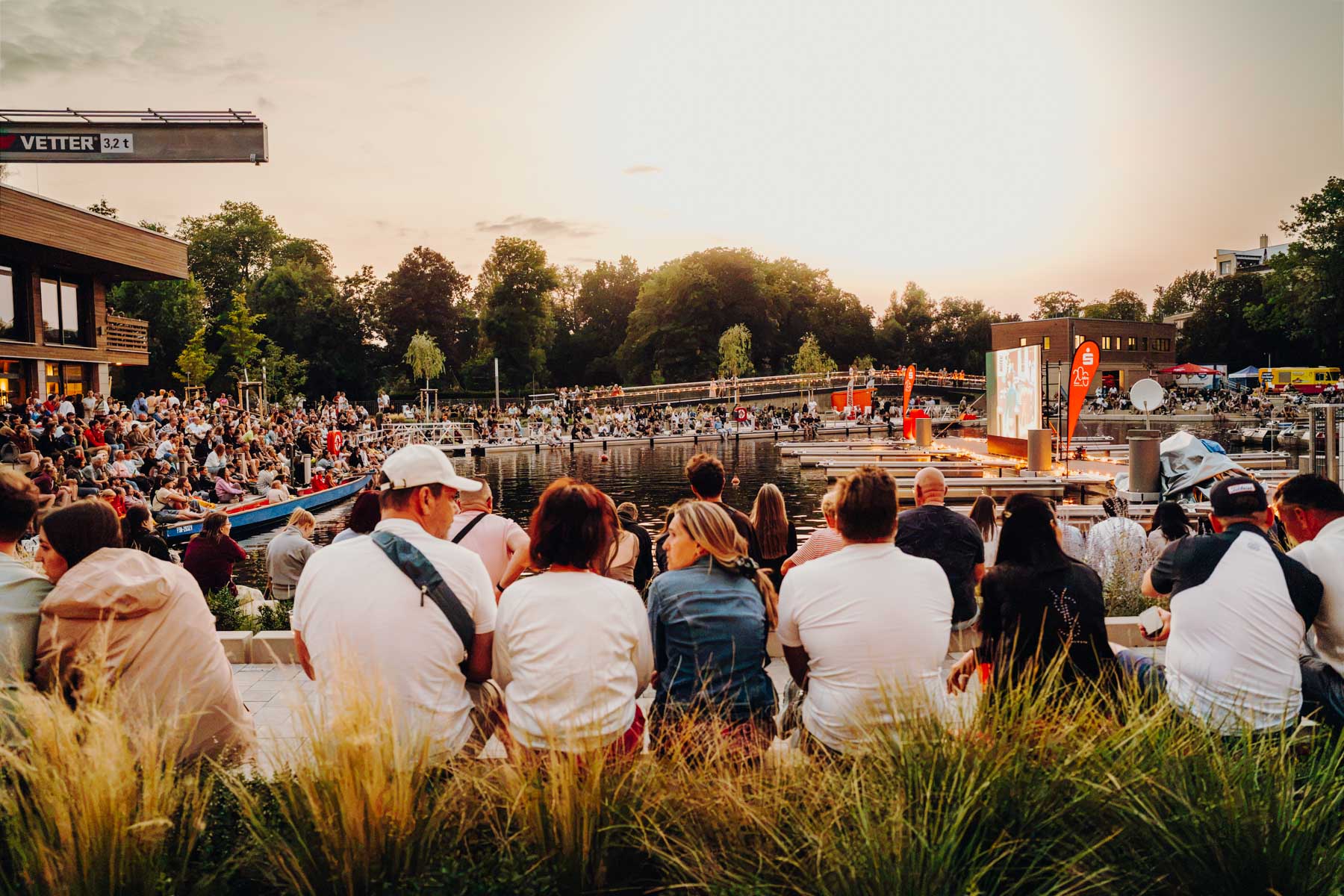 Eventfotograf Leipzig Hafenfest Eine große Menschenmenge sitzt bei Sonnenuntergang an einer Uferpromenade und schaut einen Film auf einer Leinwand am Stadthafen Leipzig