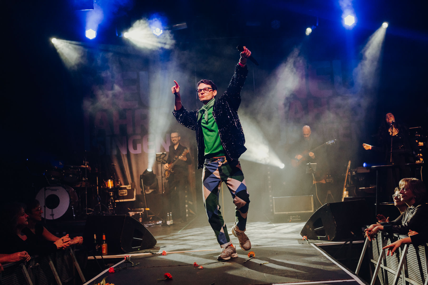 Eventfotograf Leipzig Konzert Neujahrssingen Sänger in Paillettenjacke auf einer Konzertbühne mit Band vor einem Banner mit der Aufschrift Neujahrssingen